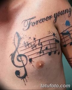 Фото тату на мужской груди: нотный стан, скрипичный ключ и надпись 'Forever Young' в черных тонах с брызгами краски.