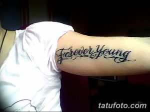 Фото тату с надписью 'Forever Young' на предплечье, выполненной изящным шрифтом. Черные чернила на светлой коже.