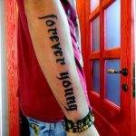 Фото тату на руке: длинная надпись 'forever young', выполненная черным шрифтом вдоль руки, с акцентом на простоту и элегантность.