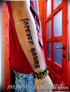 Фото тату на руке: длинная надпись 'forever young', выполненная черным шрифтом вдоль руки, с акцентом на простоту и элегантность.