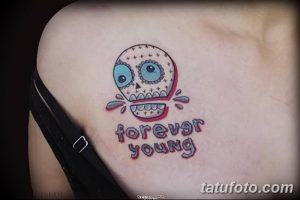 Фото тату на плече: яркий рисунок черепа с бирюзовыми глазами и надписью 'forever young', выполненный в стиле олд скул.