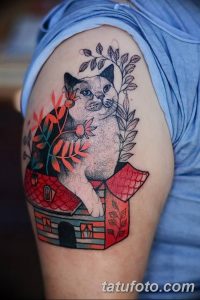 Фото татуировки на плече. Изображение кота, сидящего на крыше домика. Нежные цвета, сказочный стиль, детализированный рисунок.