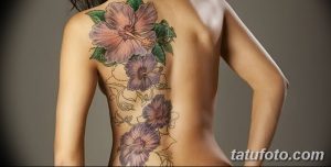 Фото тату: татуировка гибискуса на спине. Цветы гибискуса в различных оттенках фиолетового и розового, с зелеными листьями.