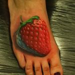 Фото тату клубники на стопе человека. Реалистичный рисунок выполнен в ярких красных и зеленых цветах, выглядит как настоящая ягода.