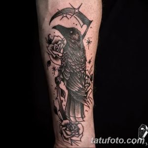 Черно-белое фото тату на предплечье с изображением ворона, держащего косу, в окружении роз и звезд. Мрачный и элегантный дизайн.