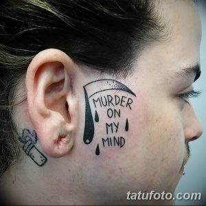 Фото татуировки за ухом: черная коса смерти с каплями и надписью 'MURDER ON MY MIND', оригинальный и провокационный дизайн.