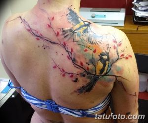 Фото акварельной татуировки птицы, сидящей на ветке цветущей сакуры на плече девушки. Нежный и романтичный рисунок татуировки.