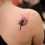 Фото минималистичной татуировки одуванчика на лопатке. Изображение выполнено в черном цвете с добавлением легких штрихов, имитирующих пух.