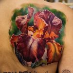 Фото тату на плече: крупный цветок ириса с яркими фиолетовыми и оранжевыми лепестками, выполненный в реалистичном стиле.