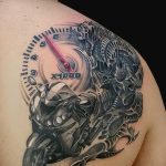 Фото тату на плече: детализированное изображение мотоцикла, объединенного с черепом и спидометром, выполненное в черно-серых тонах. Идеально для байкеров.