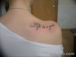 Фото минималистичной татуировки с надписью "Life is a game" на плече девушки. Черно-белый рисунок татуировки, выполненный каллиграфическим шрифтом.