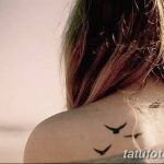 Фото тату: три маленькие черные птицы, летящие вверх, расположены на лопатке девушки. Изящный рисунок татуировки в минималистичном стиле.