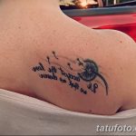 Фото татуировки одуванчика с летящими семенами и надписью на лопатке. Тату выполнено в минималистичном стиле черными чернилами.