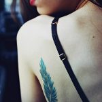 Фото тату: реалистичное изображение голубого пера на женской лопатке. Изящный рисунок татуировки, выполненный в технике акварели.