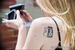 Фото тату: реалистичное изображение старинной камеры Polaroid на плече девушки. Черно-белый рисунок татуировки с высокой детализацией.