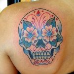 Фото тату: красочный череп в стиле sugar skull, украшенный цветами и узорами, расположен на лопатке. Яркий и оригинальный рисунок.