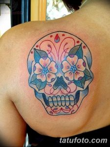 Фото тату: красочный череп в стиле sugar skull, украшенный цветами и узорами, расположен на лопатке. Яркий и оригинальный рисунок.