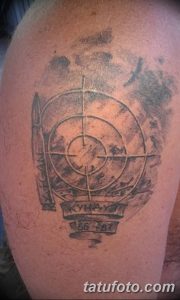 Фотография татуировки на плече, выполненной в темных тонах. Изображение включает в себя прицел, оружие и надпись, создавая милитаристический образ.