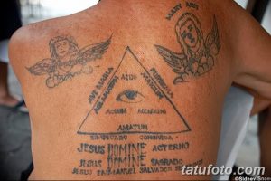 Фото тату на спине: два ангела, фланкирующие треугольник с глазом. Религиозный мотив с использованием символики и надписей. Черно-серый рисунок.