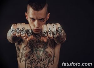 Фото мужчины с множеством татуировок на теле, включая черепа на пальцах и сложные геометрические узоры. Рисунок татуировки выполнен в тёмных тонах, создавая мрачный и загадочный образ.