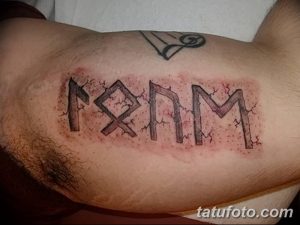 Фото тату на бицепсе с изображением рунической надписи, выполненное в черном цвете с эффектом трещин и потертостей.