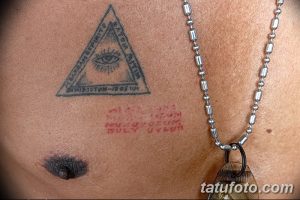 Фото тату на груди: изображение Всевидящего ока в треугольнике и надпись на английском языке. Рисунок татуировки в синих и красных тонах.