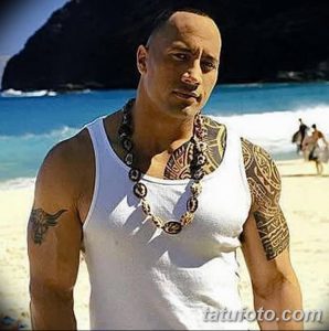 Фотография Двейна Джонсона на пляже с татуировкой на плече и груди, в белой майке и с ожерельем из бусин.