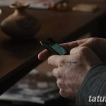 Фото тату: небольшой черный крест на тыльной стороне руки мужчины, держащего телефон. Минималистичный рисунок татуировки.