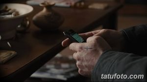 Фото тату: небольшой черный крест на тыльной стороне руки мужчины, держащего телефон. Минималистичный рисунок татуировки.