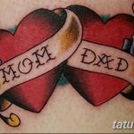 Фото татуировки двух сердец с надписями 'Mom' и 'Dad' на лентах, украшенных цветами. Цветной рисунок татуировки.
