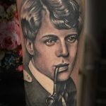 Фотография реалистичной татуировки на руке с изображением Сергея Есенина, курящего трубку. Детализированный рисунок с использованием оттенков серого и красных акцентов.
