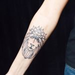 Фото тату на предплечье: минималистичное изображение головы льва с короной, выполненное тонкими линиями. Черно-белая татуировка в лаконичном стиле.