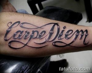 Фото татуировки 'Carpe Diem' на предплечье, выполненной красивым каллиграфическим шрифтом. Черно-белый рисунок с плавными линиями и изящными завитушками.
