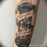 Фото тату: реалистичные часы с песком, обвитые лентой с надписью 'Carpe Diem' на предплечье мужчины. Черно-серые тона, детальная проработка.