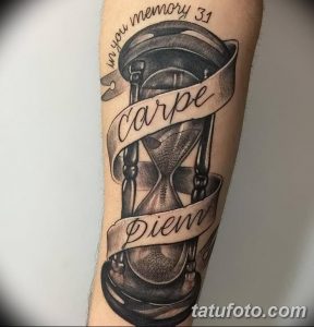 Фото тату: реалистичные часы с песком, обвитые лентой с надписью 'Carpe Diem' на предплечье мужчины. Черно-серые тона, детальная проработка.