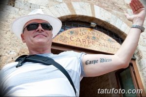 Фото тату надписи 'Carpe Diem' выполненной на руке на фоне архитектурного элемента. Черно-белая татуировка, реалистичный стиль, интересный фон.