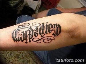 Фото тату надписи 'Carpe Diem' выполненной готическим шрифтом на предплечье. Черно-белая татуировка, четкие линии, классический стиль.