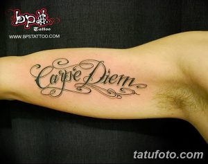 Фото тату надписи 'Carpe Diem' выполненной изящным шрифтом с завитушками на руке. Черно-серая татуировка, тонкие линии, элегантный дизайн.