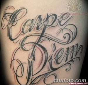 Фото тату надписи 'Carpe Diem' черным цветом, выполненной каллиграфическим шрифтом с декоративными завитушками на руке.
