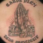 Фото тату 'Carpe Diem' над изображением молящихся рук на груди. Черно-серая татуировка с религиозным подтекстом и детальной проработкой.