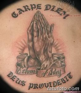Фото тату 'Carpe Diem' над изображением молящихся рук на груди. Черно-серая татуировка с религиозным подтекстом и детальной проработкой.