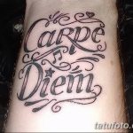 Фото тату надписи 'Carpe Diem' с витиеватыми узорами, сердечком и звездочкой на запястье. Черно-серая татуировка с изящным дизайном.