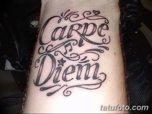 Фото тату надписи 'Carpe Diem' с витиеватыми узорами, сердечком и звездочкой на запястье. Черно-серая татуировка с изящным дизайном.