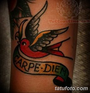 Фото тату 'Carpe Diem' в виде ленты с ласточкой в стиле олдскул на предплечье. Яркие цвета, четкие контуры и традиционный дизайн.