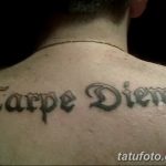 Фото тату надписи 'Carpe Diem' готическим шрифтом на спине, между лопатками. Черно-серая татуировка с выразительными линиями и завитками.