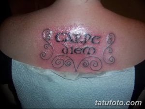 Фото тату надписи 'Carpe Diem' на спине, обрамленной декоративными завитками. Черно-серый рисунок, женственный дизайн. Подходит для тех, кто ценит красоту и элегантность.