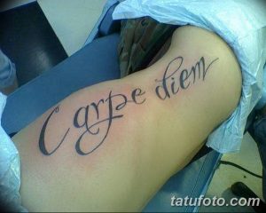 Фото тату надписи 'Carpe Diem' на плече. Черный контур, каллиграфический шрифт, плавные линии. Подходит для тех, кто ценит красоту и гармонию.