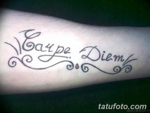 Фото тату надписи 'Carpe Diem' с декоративными завитками и точками. Черно-серый рисунок, выполненный на руке. Винтажный стиль, подходит для любителей классики.