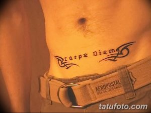 Фото тату надписи 'Carpe Diem' на боку мужчины. Черный рисунок татуировки с декоративными элементами, выполненный в стиле минимализм.