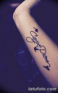 Фото тату с надписью 'Carpe Diem', украшенной небольшими изображениями якоря и стрелы. Минималистичный рисунок татуировки на руке.